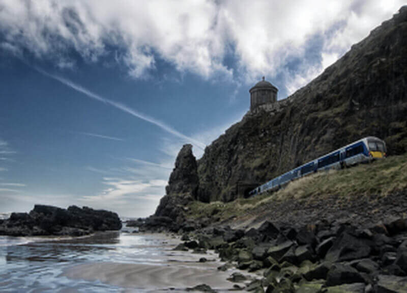 Scenic coastal train passing rugged cliffs and shoreline, offering serene views of distant hills under a bright blue sky.