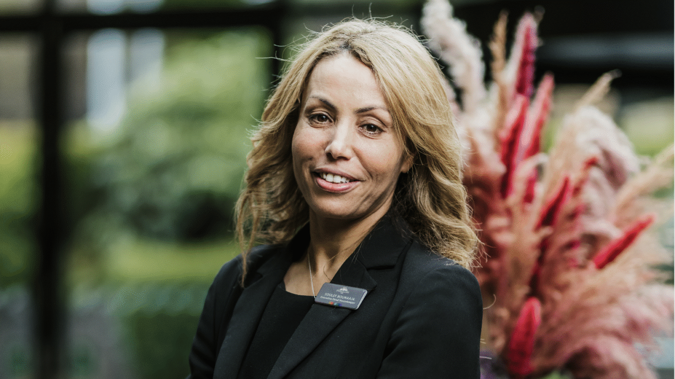 Friendly hotel staff member in smart attire welcomes guests with a smile, standing by decorative pampas grasses in a lush setting.