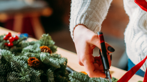 Person crafting festive wreath with pine branches and red ribbon on wooden table, showcasing hotel holiday workshop.