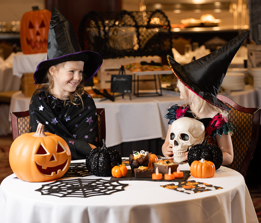 Children in witch hats enjoy Halloween treats at themed dining table, featuring pumpkins and spooky decor.