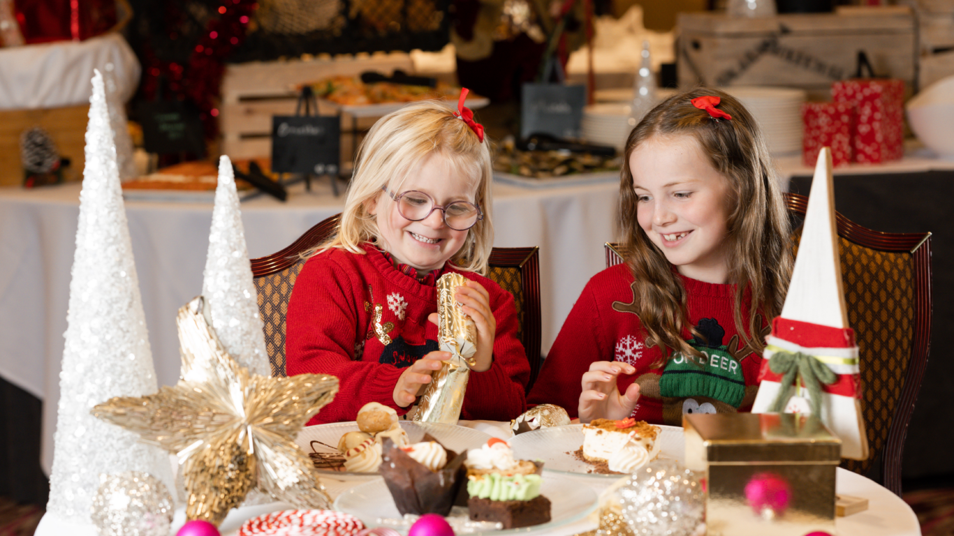 Children in festive jumpers enjoying dessert at Christmas-themed hotel table with decorations and pastries.
