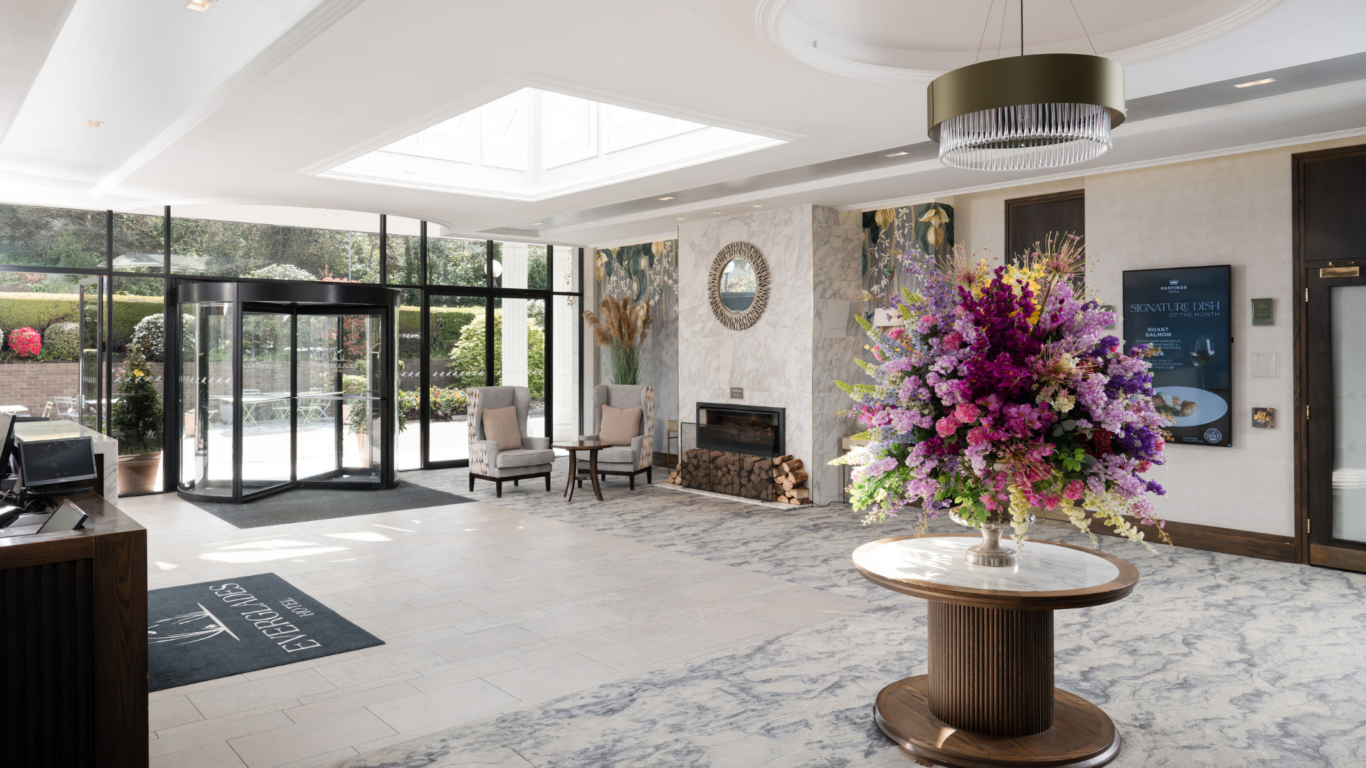 Elegant hotel lobby with bright skylight, revolving door, fireplace, plush seating, and vibrant floral centrepiece.