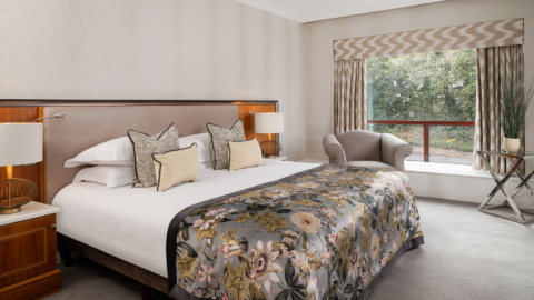 Elegant room with king bed, floral quilt, cosy seating and large window overlooking lush greenery. Ideal for a serene getaway.