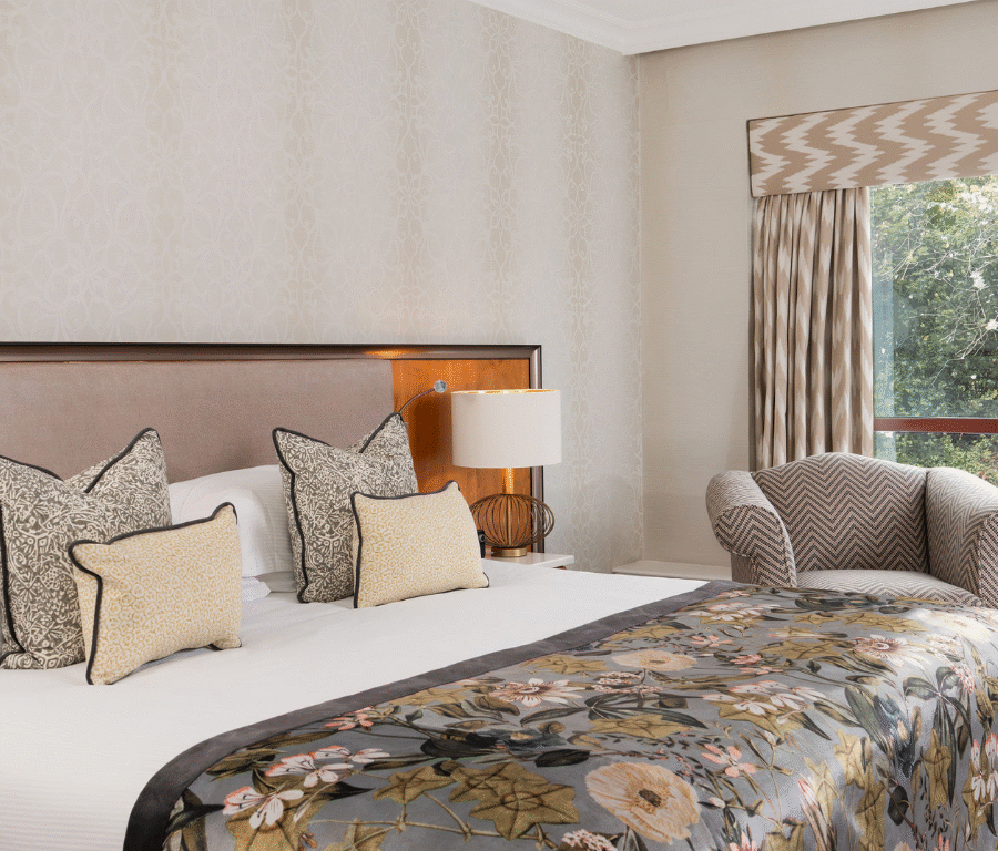 Elegant hotel room with plush king bed, floral duvet, cushioned armchair, large window view, and neutral d&eacute;cor.
