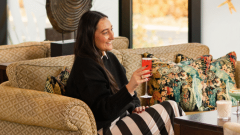 Guest relaxing in stylish lounge with vibrant décor, enjoying cocktails by large windows overlooking serene autumn scenery.